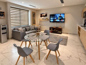 A modern living room with a sofa, a glass coffee table, and two chairs. There's a television mounted on the wall, a bookshelf, and a kitchen area with cabinets.
