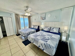 A hotel room with two beds, a ceiling fan, a view of the ocean, and a balcony.