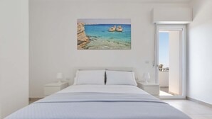 A bedroom with a bed, white nightstands, a painting of a coastal scene, and a door leading to an outdoor area. - 3099 Residence Marelena- Bilo Grecale by Barbarhouse (Squinzano)