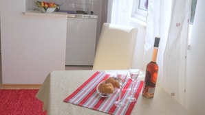 A small kitchen with white cabinets, a window with sheer curtains, and a dining area with a table set for two. - Apartments Franka (Dubrovnik)