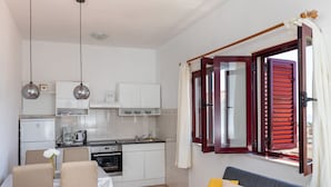 A compact kitchen with white cabinets, a dining table with chairs, a sofa with cushions, and a window with red shutters. - Apartments Franka (Dubrovnik)