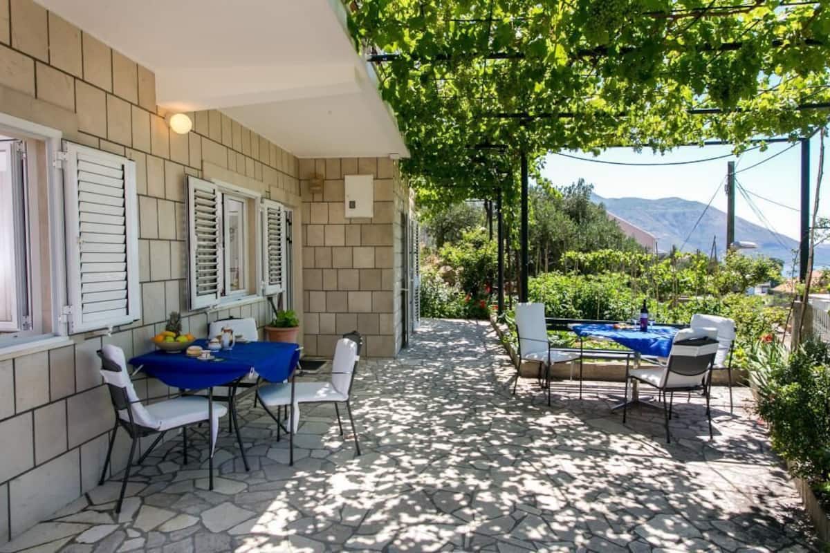 An outdoor patio with stone flooring, white chairs, a blue tablecloth-covered table, and a view of mountains in the background.