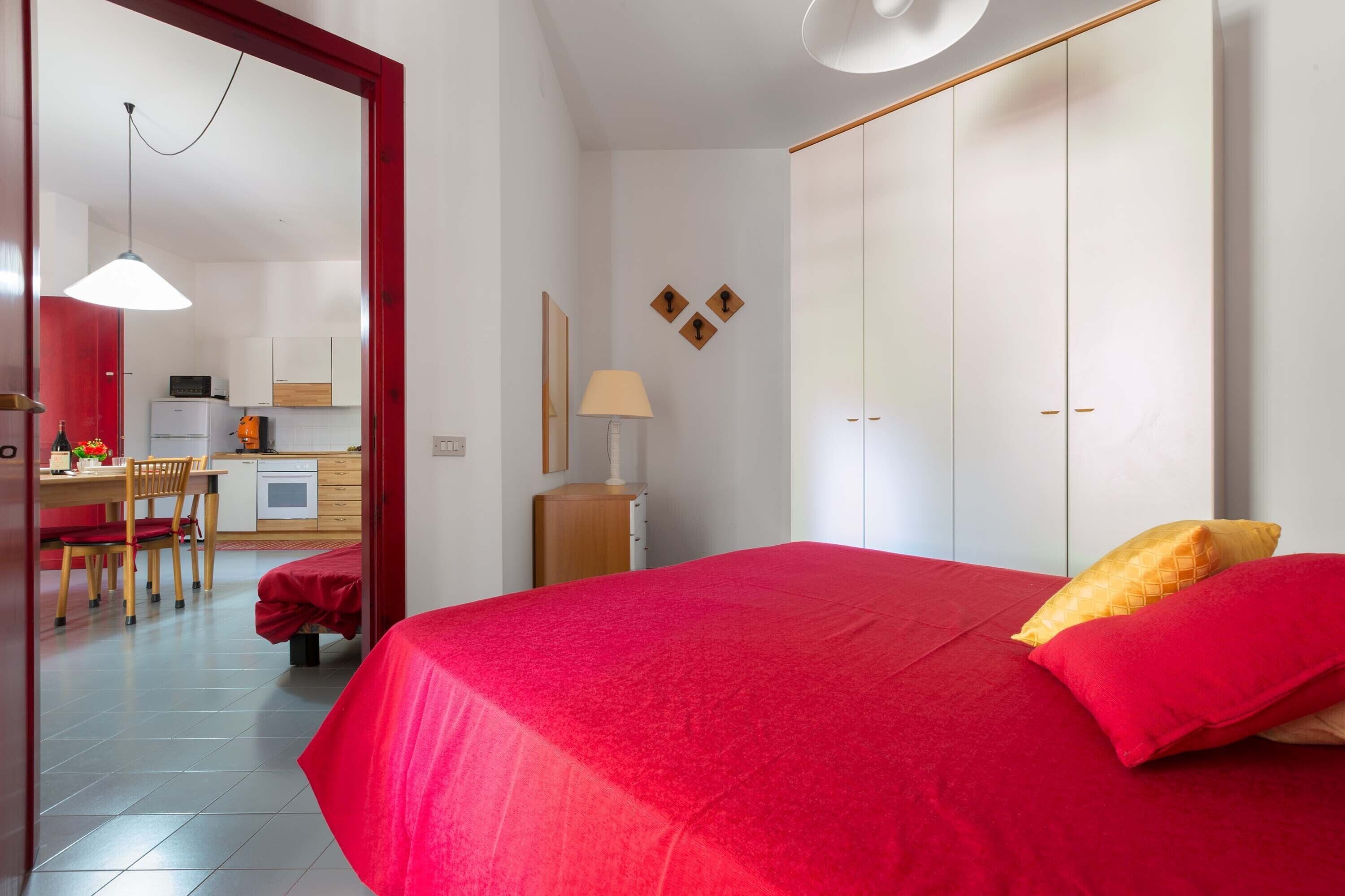 A bedroom with a red bedspread, a wooden nightstand with a lamp, and a wardrobe.