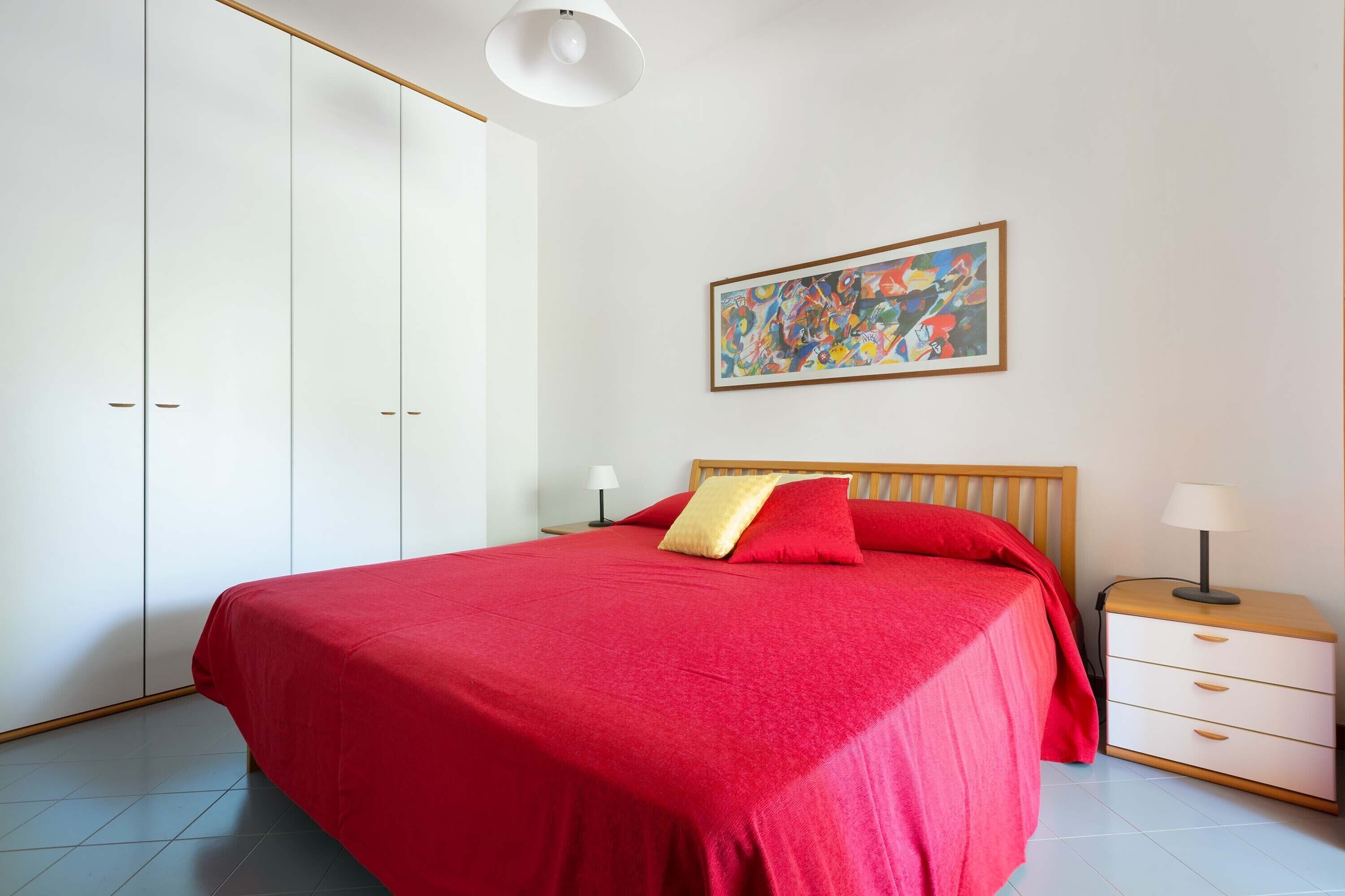 A bedroom with a large bed, a nightstand with a lamp, a wardrobe, and a colorful abstract painting on the wall.