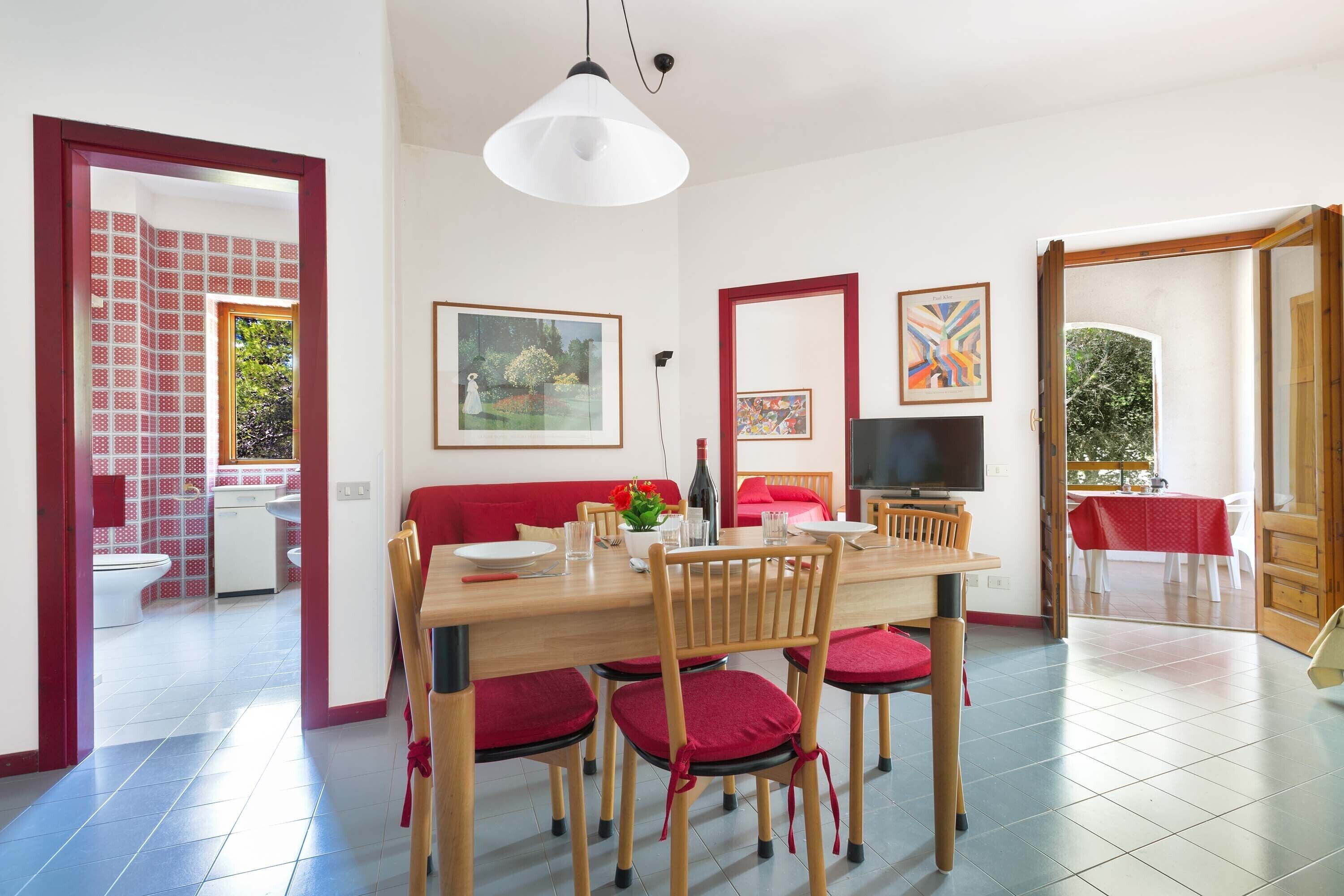 A dining area with a wooden table, chairs, and a television. There are framed pictures on the wall, a red sofa, and a red tablecloth on a table in the background.