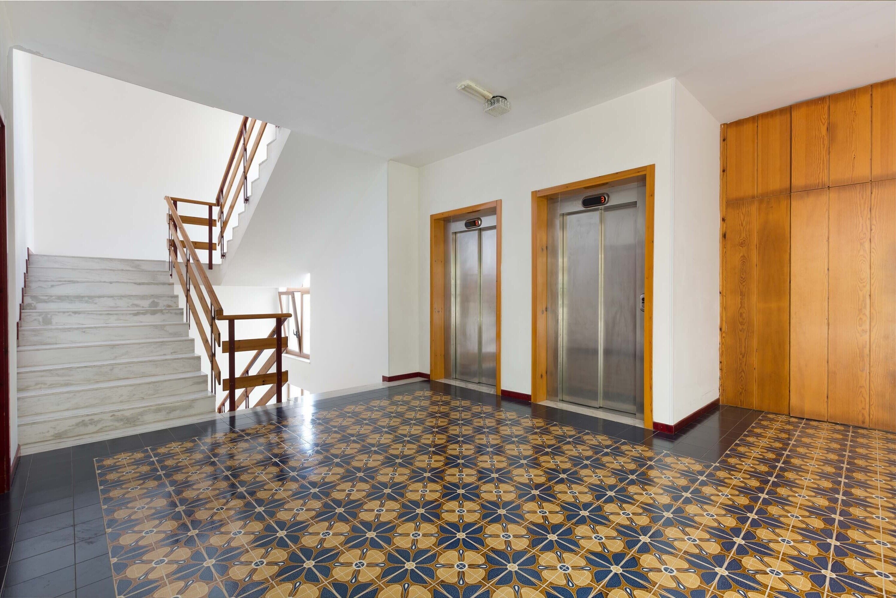 Un pasillo amplio con piso estampado, una escalera de madera y dos ascensores.