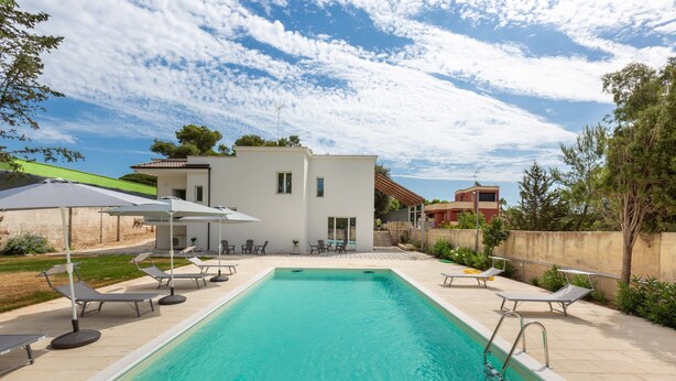 A rectangular swimming pool with lounge chairs and umbrellas, surrounded by a paved patio and a modern building. - 2488 Villa Gaia by Perle di Puglia (Cavallino)