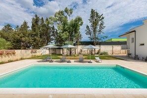 A rectangular swimming pool with clear blue water, surrounded by lounge chairs and umbrellas, with a backdrop of trees and a building. - 2488 Villa Gaia by Perle di Puglia (Cavallino)