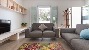 A modern living room with two grey sofas, a colorful rug, a wooden shelf with decorative items, and a large window with a view of trees. - 2488 Villa Gaia by Perle di Puglia (Cavallino)