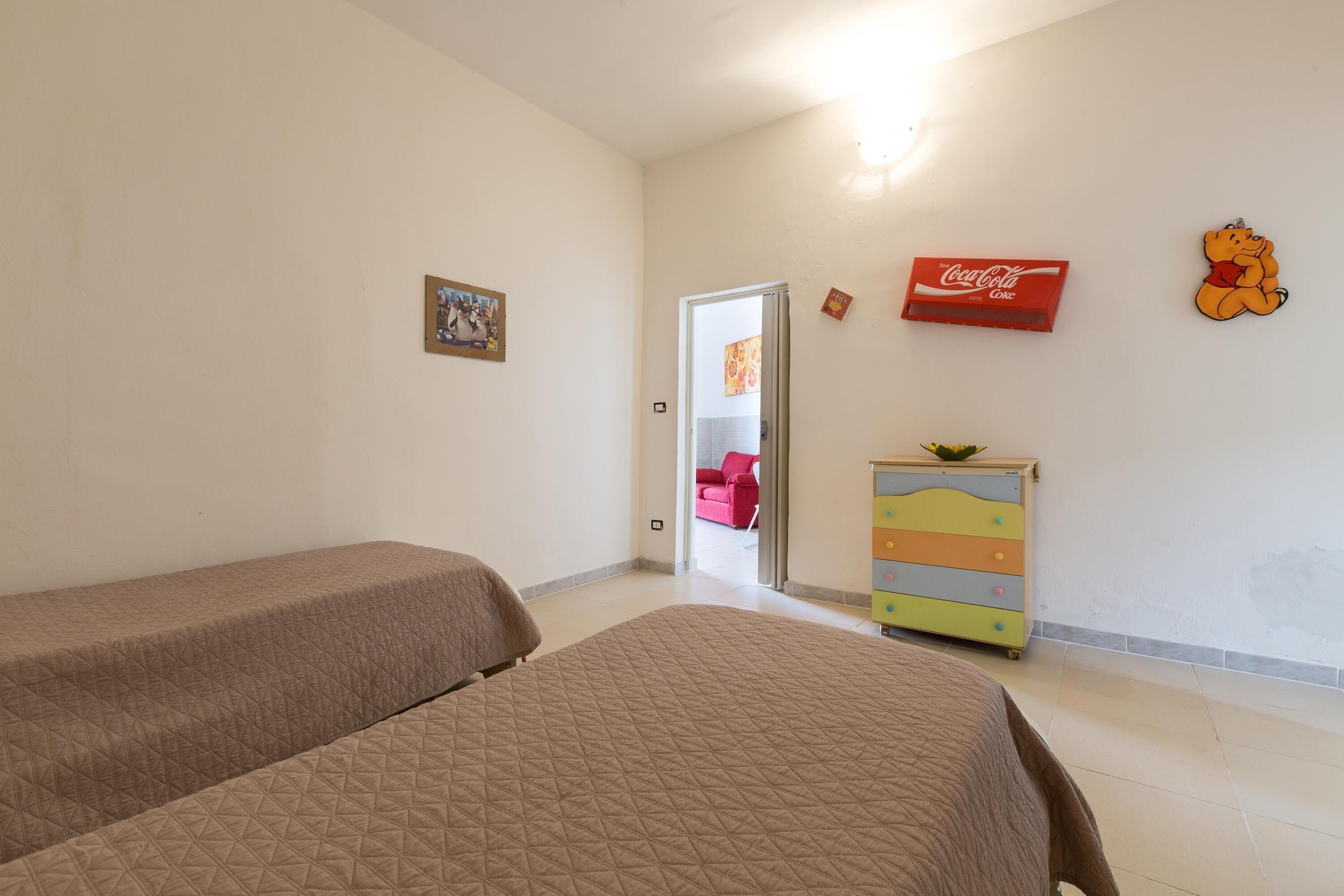 A room with two beds, a colorful dresser, a red chair, and a door leading to another room.