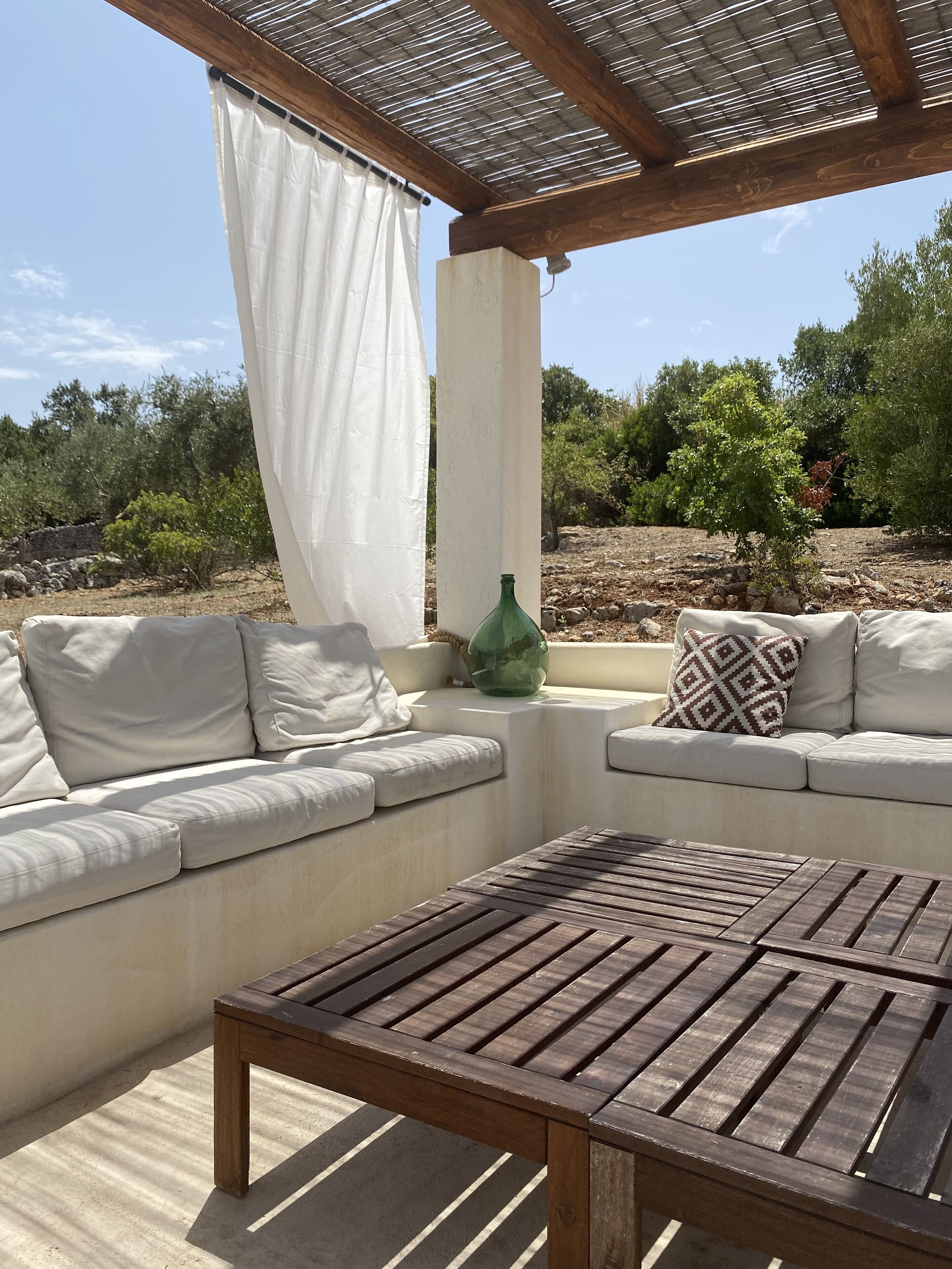 An outdoor seating area with a wooden table and white cushioned sofas, a pergola overhead, and a view of a rocky landscape with trees.