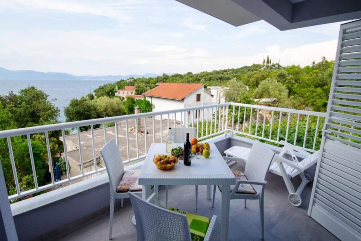 A balcony with a table set for two, a bottle, and a bowl of fruit.