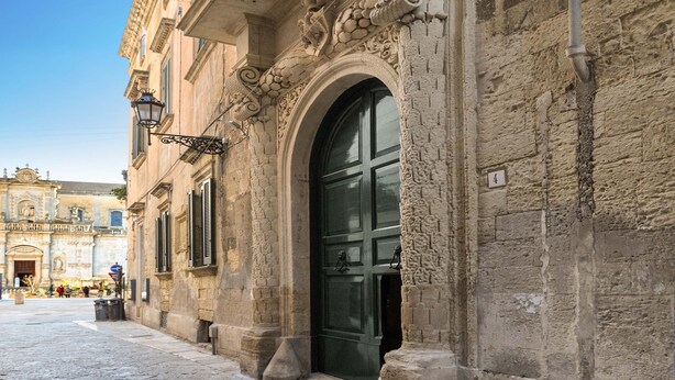 A historic building with ornate stonework, a large arched doorway, and a street view with other buildings in the background. - 2661 Porta del Duomo Luxury by Barbarhouse (Lecce)