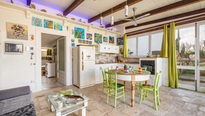 A spacious kitchen with a dining area, featuring a stone floor, wooden beams, and a large window with curtains.