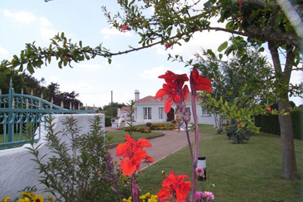 A garden with a white building, a blue bridge, and a tree with red flowers. - Chez Bernadou (Machecoul-Saint-Même)