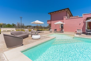 Uma área de piscina com espreguiçadeiras e guarda-sóis, um edifício rosa e um céu azul límpido.