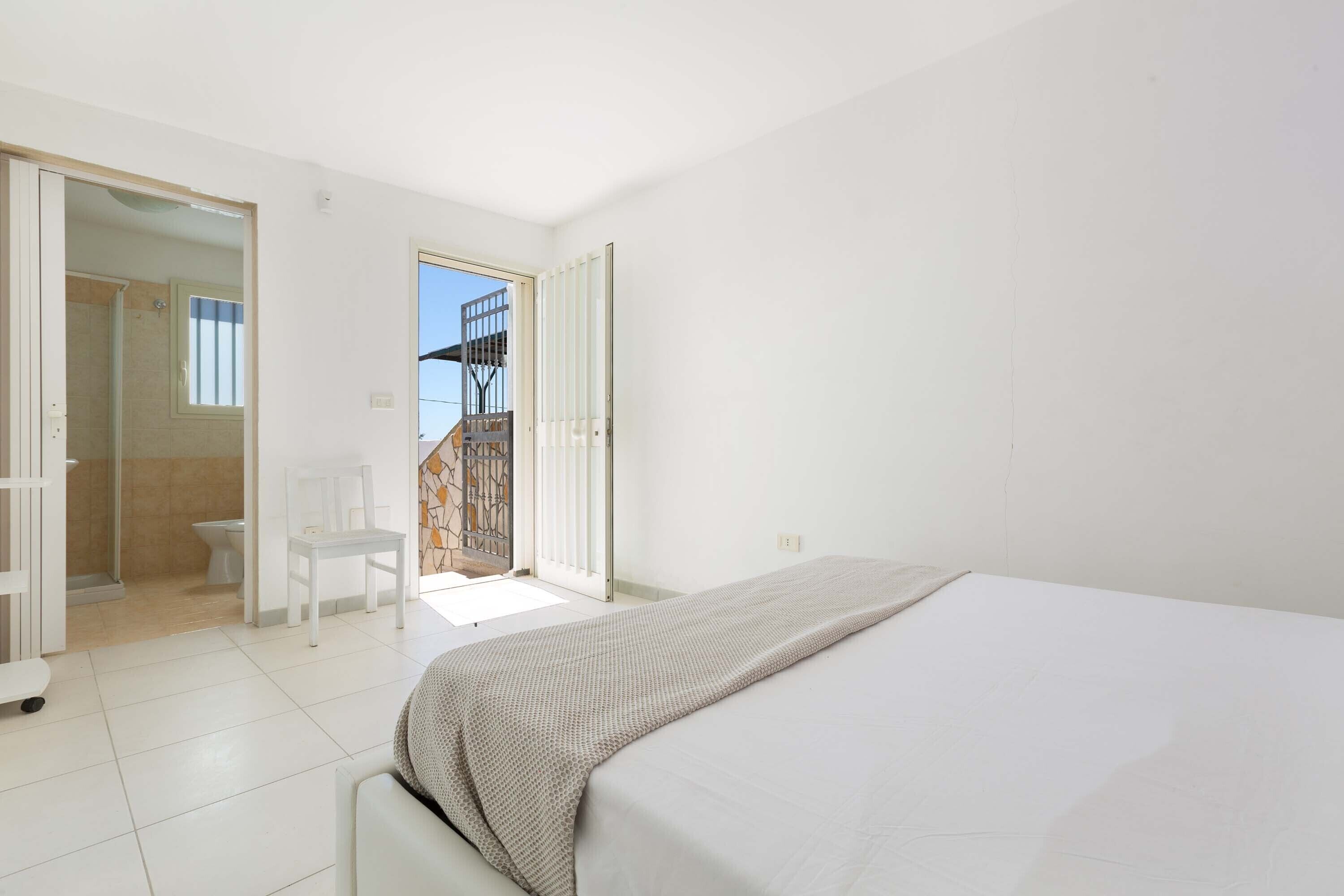 Un dormitorio con cama, una silla y un baño a la vista a través de una puerta abierta.