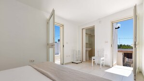 Ein Schlafzimmer mit einem Bett, einem weißen Stuhl und einem kleinen Tisch. Ein Badezimmer ist durch eine offene Tür sichtbar.