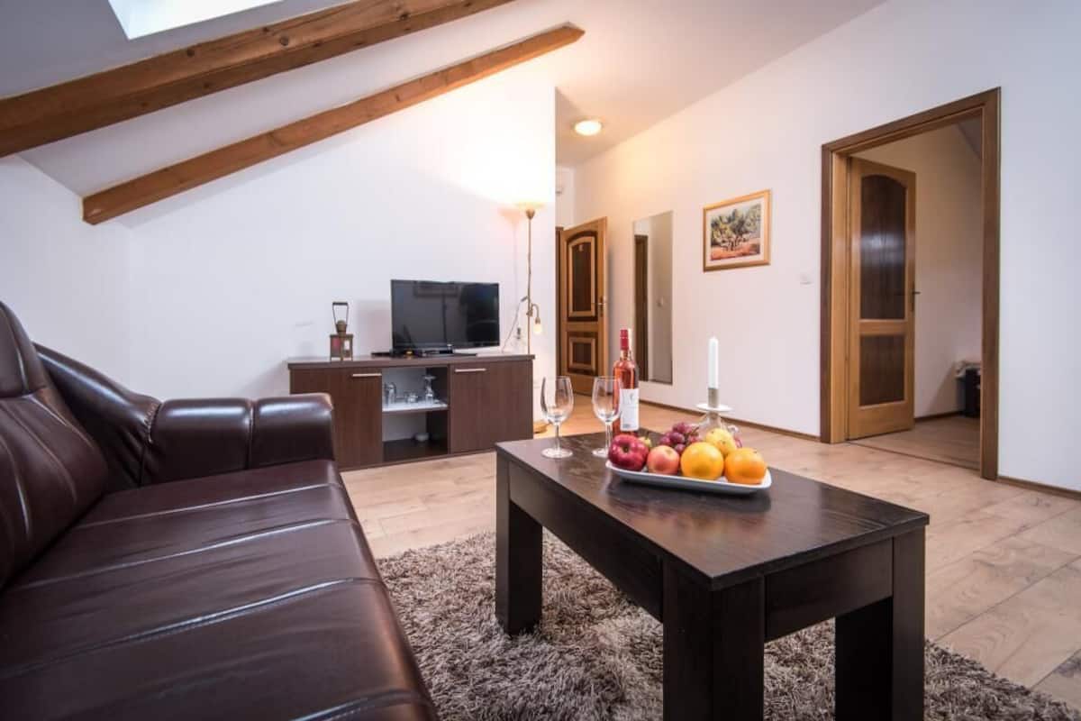 A modern living room with a leather sofa, a wooden coffee table with fruit, a TV, and a kitchen area in the background.