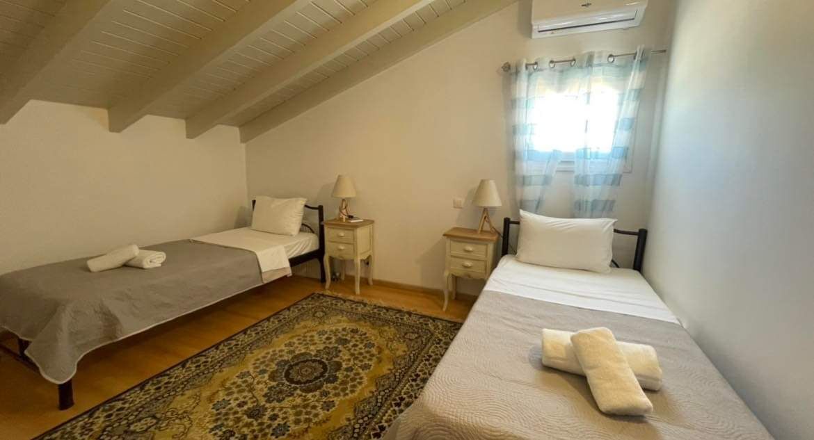 A small, cozy room with two single beds, a wooden ceiling, a patterned rug, and a window with curtains.