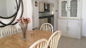 Un comedor con una mesa de madera, sillas blancas, un jarrón con flores secas y un aparador blanco con puertas de cristal.