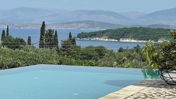 Una piscina azul cristalina con borde de piedra, rodeada de vegetación y con vistas a un paisaje costero con montañas al fondo.