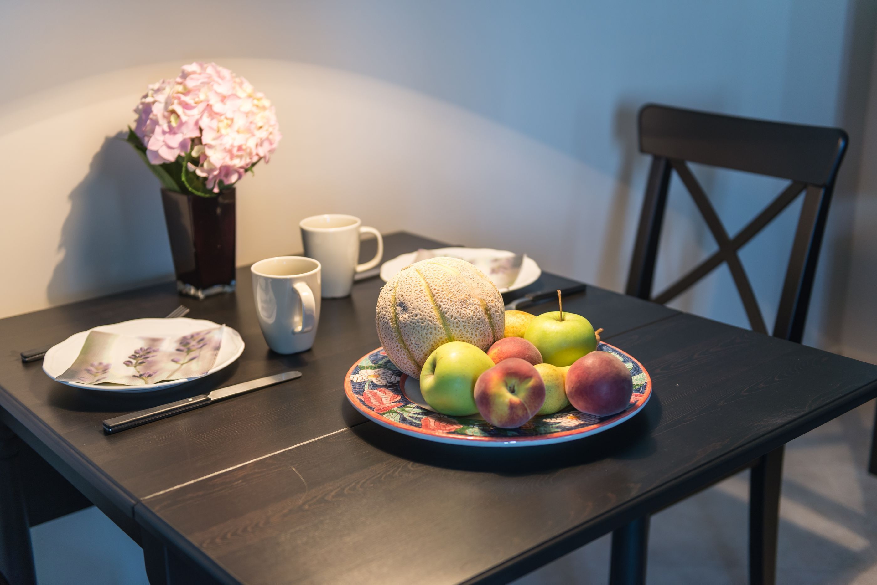Una mesa de comedor con un plato de frutas, un melón y un jarrón con flores.