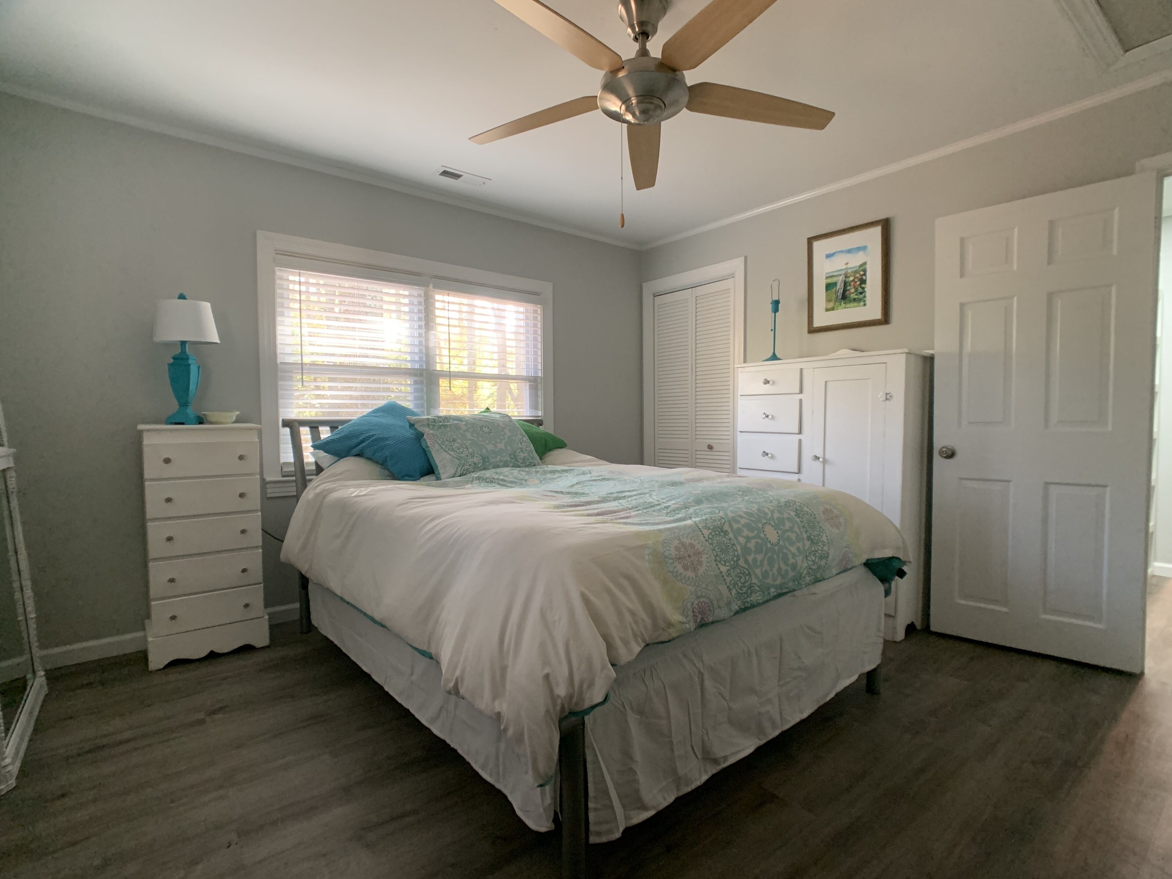 A bedroom with a bed, a ceiling fan, a window with blinds, a dresser, and a door.