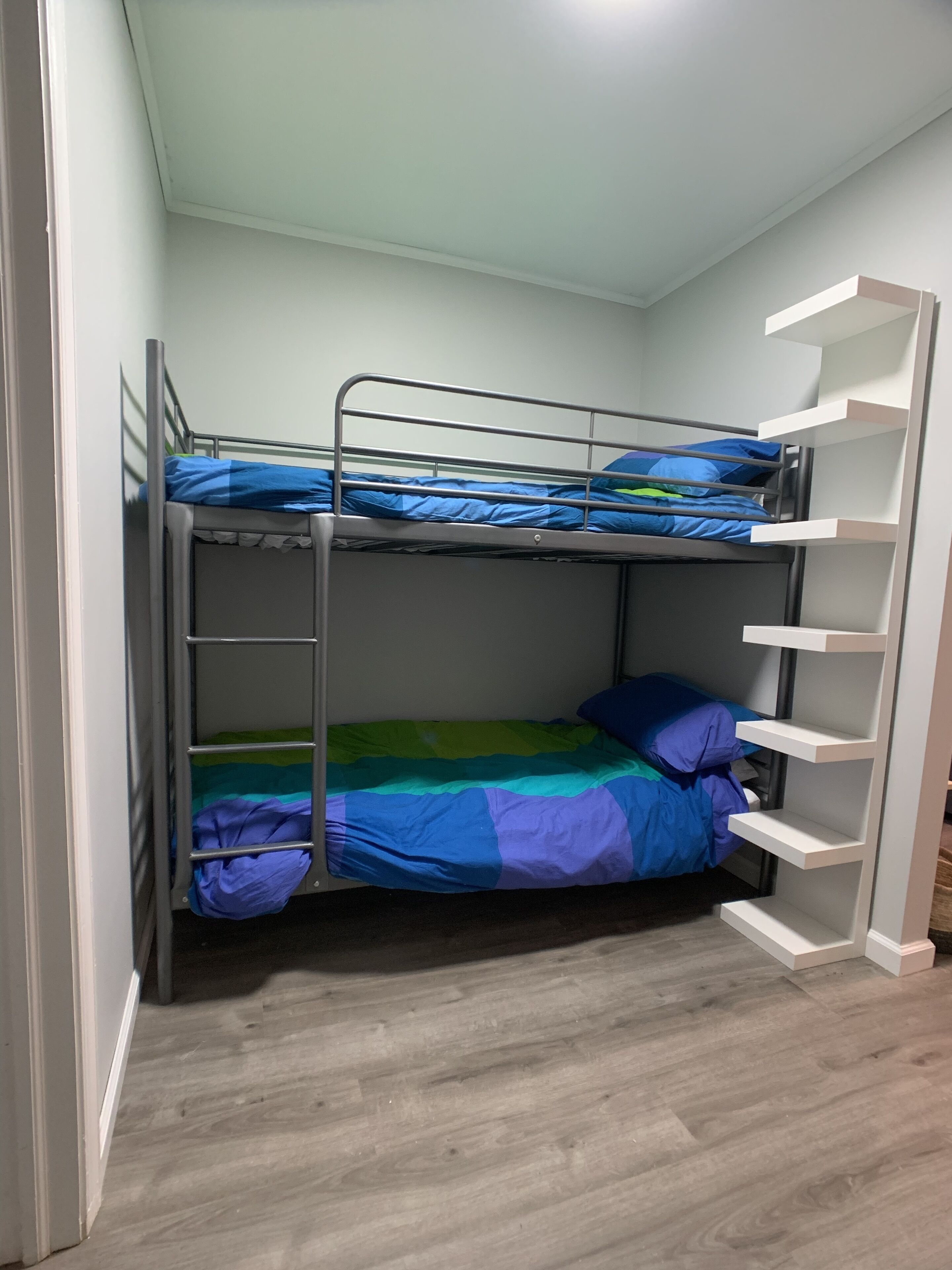 Een stapelbed met een metalen frame, een onderste bed met een kleurrijke deken, en een witte plankkast aan de rechterkant.
