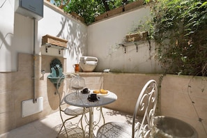 A small outdoor patio with a round metal table, two chairs, a decorative urn, and a small shelf with baskets.