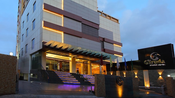 A modern hotel with a curved glass facade, multiple floors, and a prominent entrance with steps and illuminated signage. - The Garuda Hotels (Thrissur)