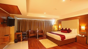 A hotel room with a large bed, a desk, a chair, a TV, and a window with curtains. - The Garuda Hotels (Thrissur)