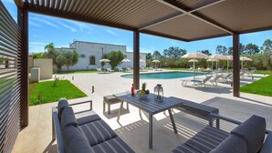 A covered outdoor area with a pool, lounge chairs, and a table with a bowl of fruit and a bottle. - 2283 Villa La Paiara by Perle di Puglia (Melendugno)