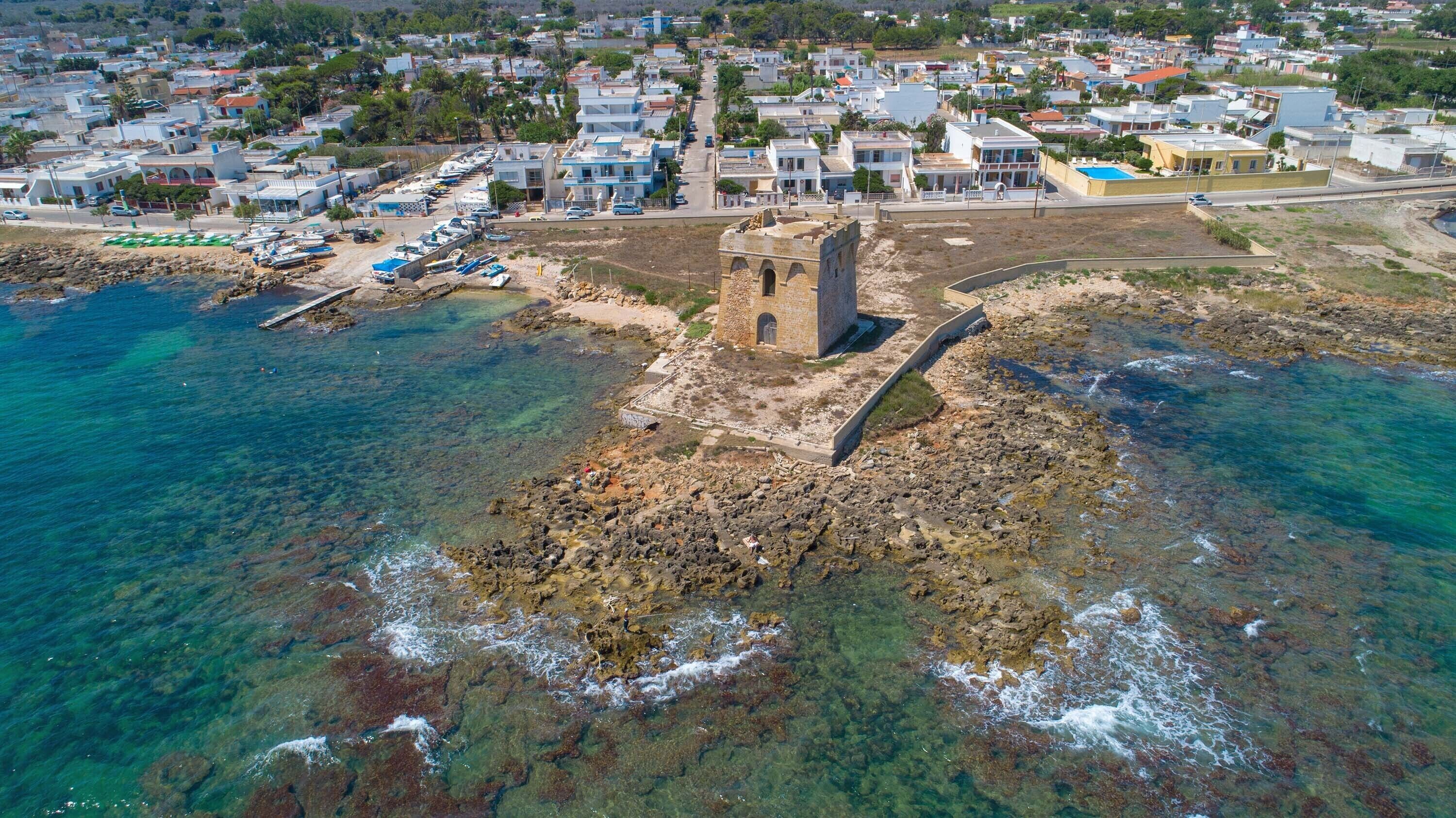 Vista aérea de un pueblo costero con una imponente torre de piedra, aguas azul cristalino y un puerto deportivo con barcos.