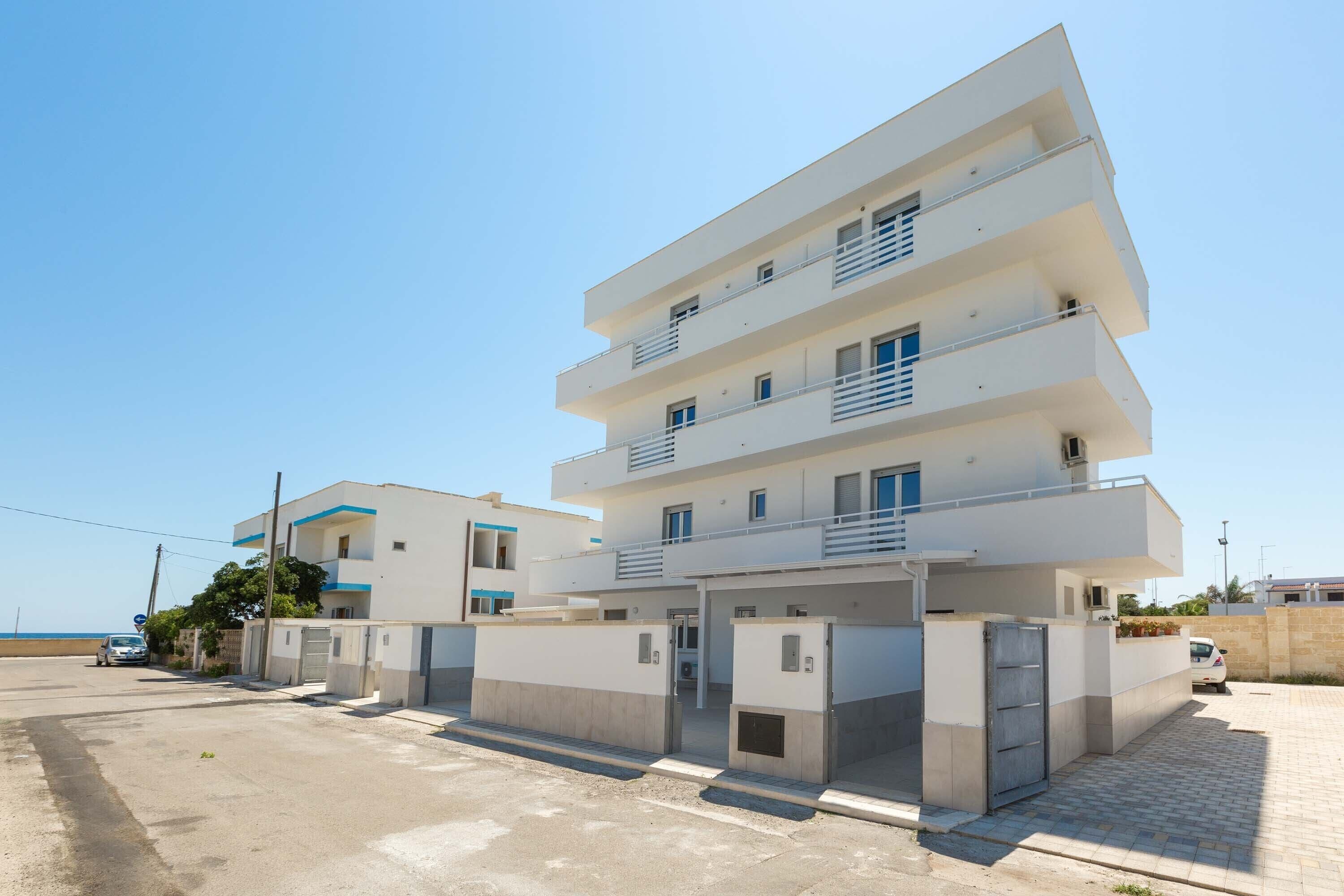 A multi-story residential building with balconies, a parking area, and a clear view of the sea.