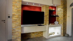 A modern living room with a large flat-screen TV mounted on a brick wall, a white entertainment unit below, and a small shelf with decorative items. - Modern and Homely Apartment in Marsaskala (Marsaskala)