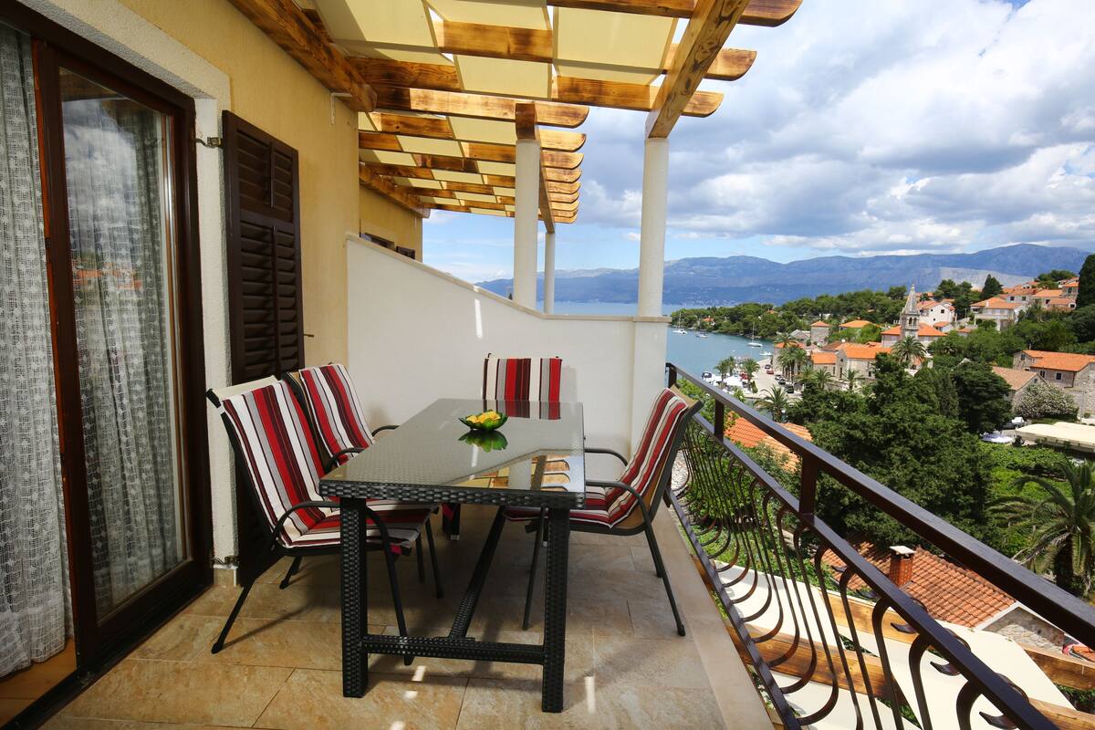 Un balcon avec une table et des chaises, offrant une vue panoramique sur une ville côtière et les montagnes.