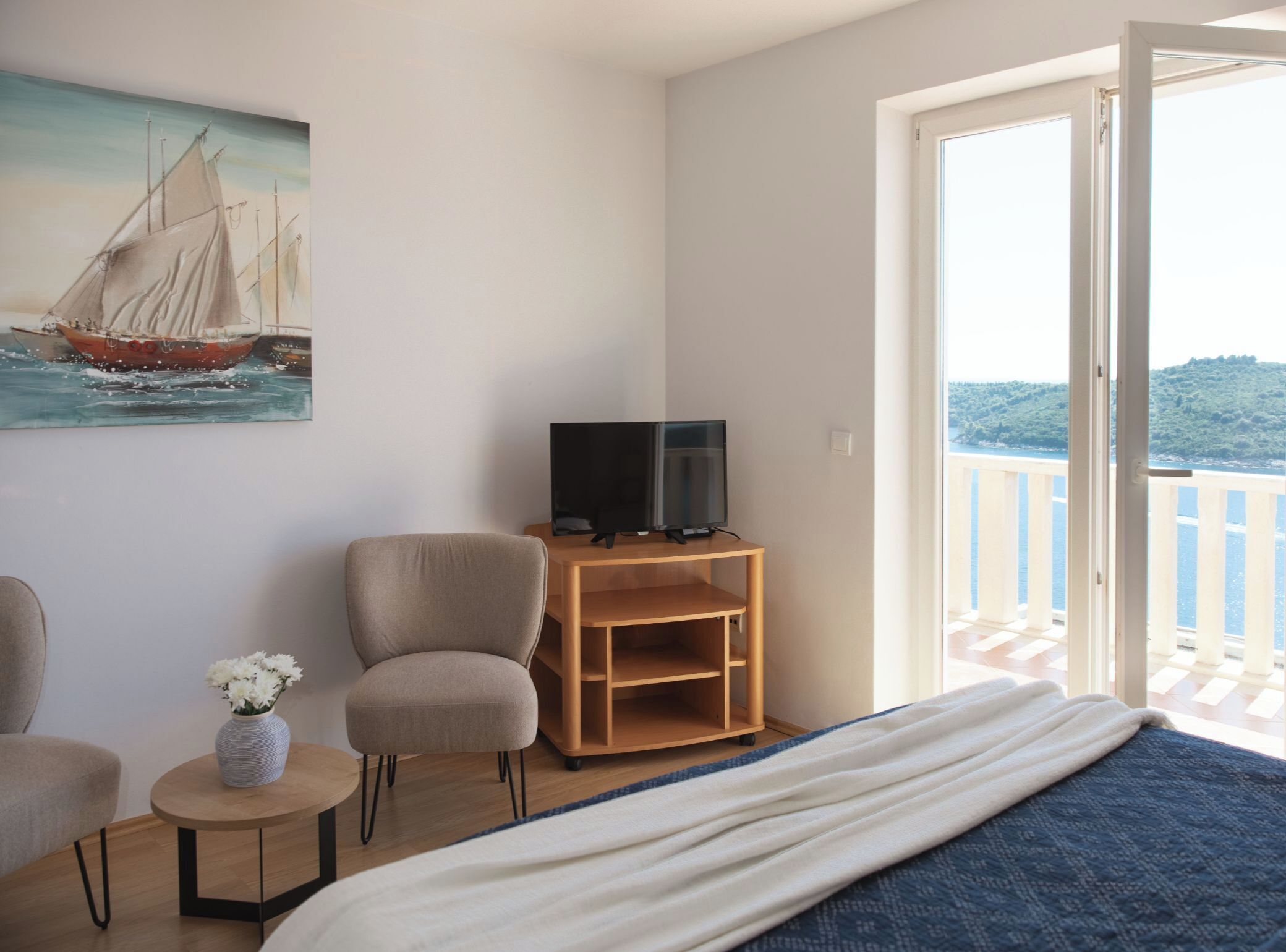 A bedroom with a bed, a TV on a wooden stand, a chair, a small table with a vase, and a painting of a sailboat.