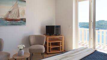 A bedroom with a bed, a TV on a wooden stand, a chair, a small table with a vase, and a painting of a sailboat.