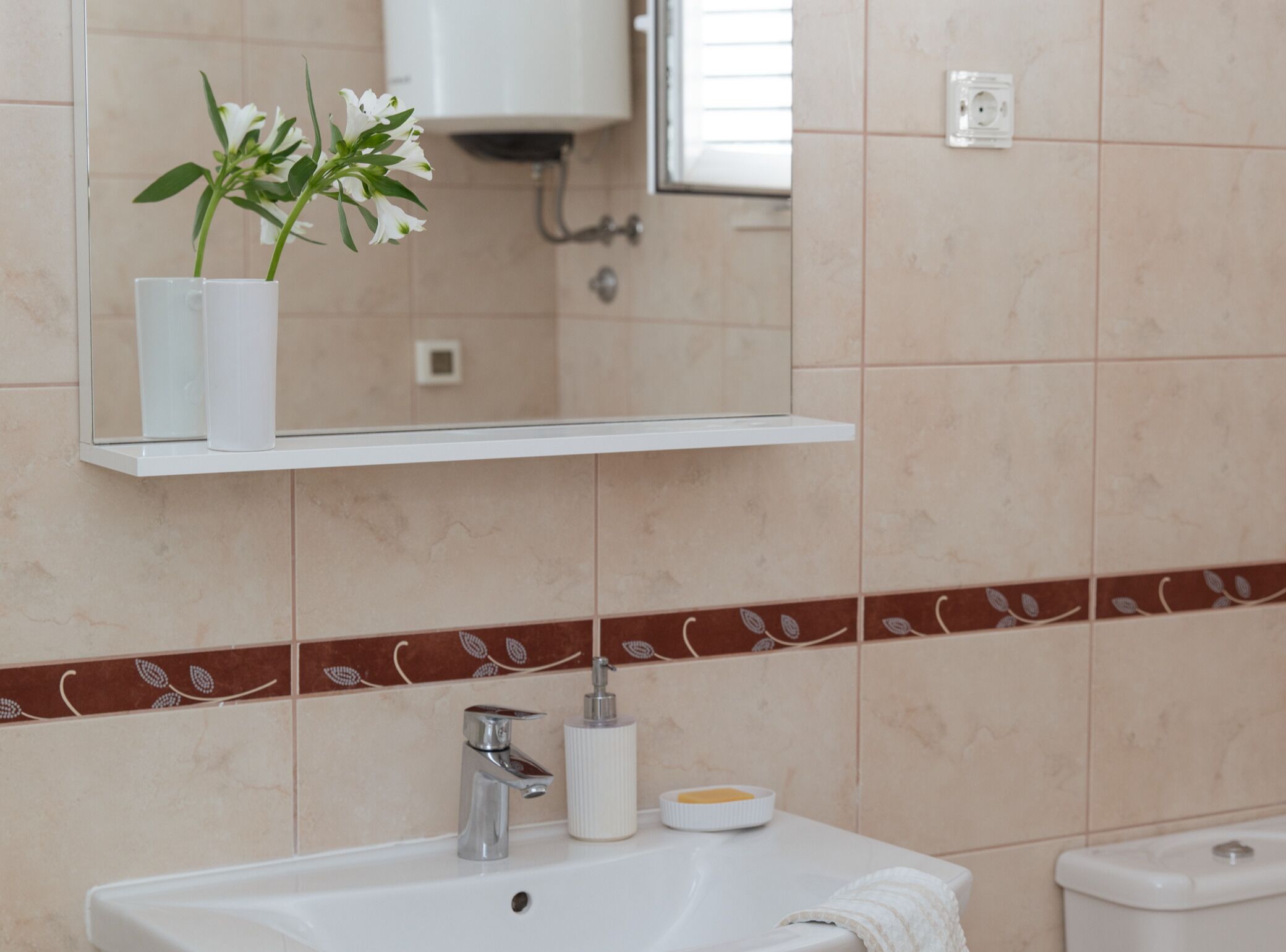 A bathroom with a white sink, a mirror above it, a white vase with flowers, and a toilet.