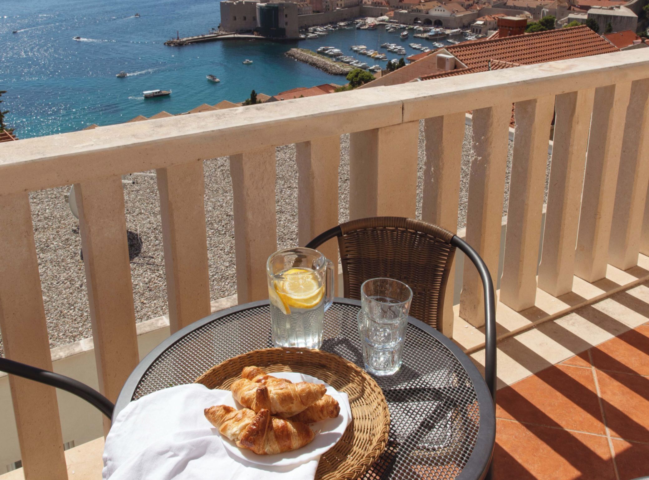 A balcony with a table set for two, featuring croissants and a glass of water, overlooking a marina with boats and historic buildings.