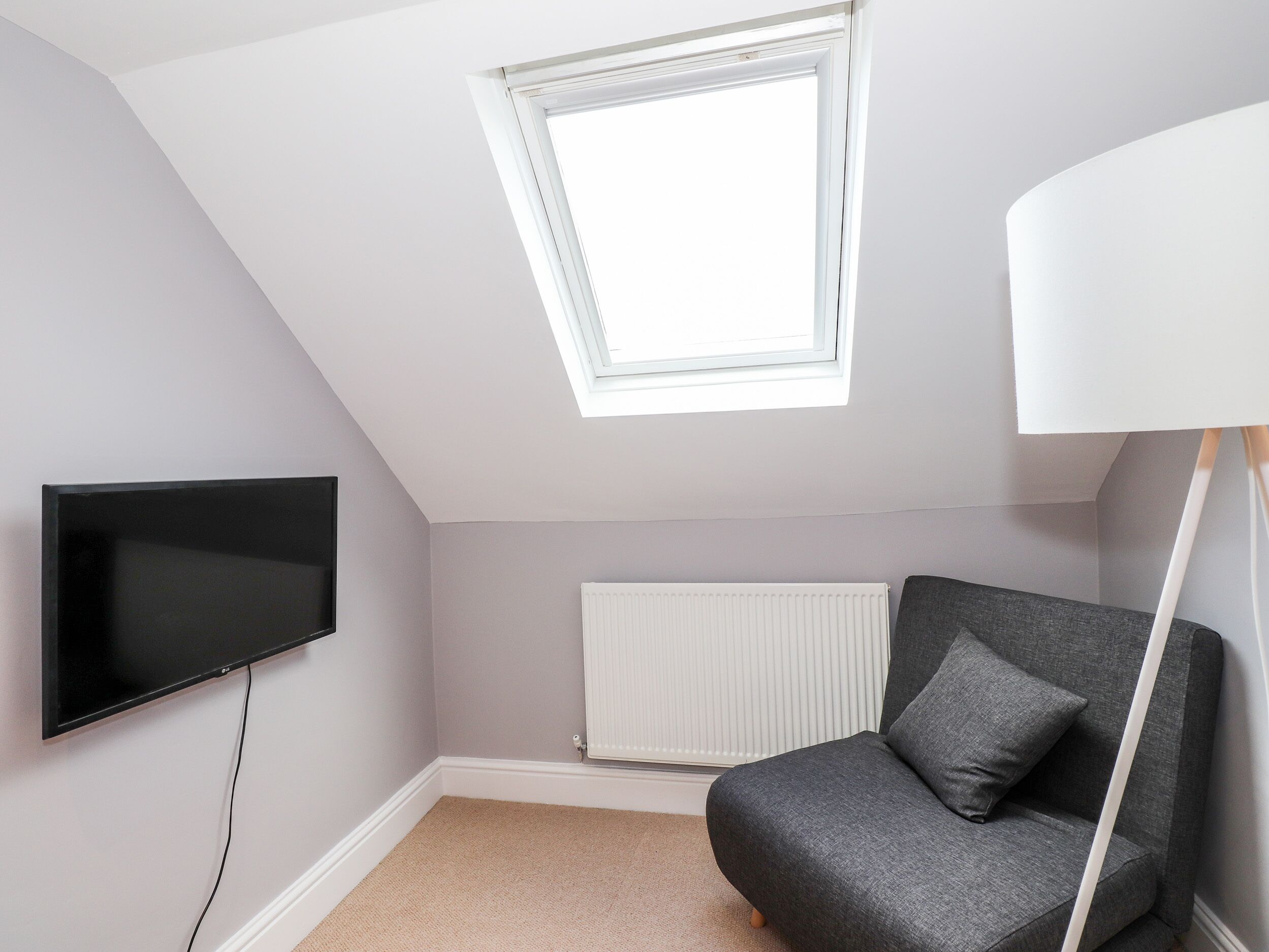Une pièce avec un plafond en pente, un téléviseur à écran plat fixé au mur, un radiateur, un canapé gris avec un coussin et une lucarne.