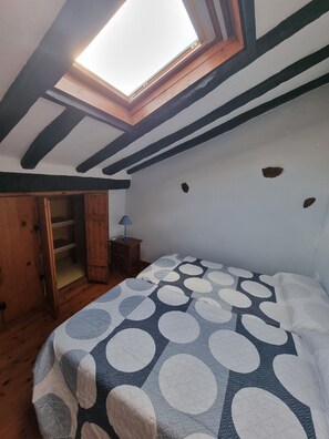 Quarto com claraboia, cama com colcha de bolinhas, guarda-roupa de madeira e mesa de cabeceira com abajur.