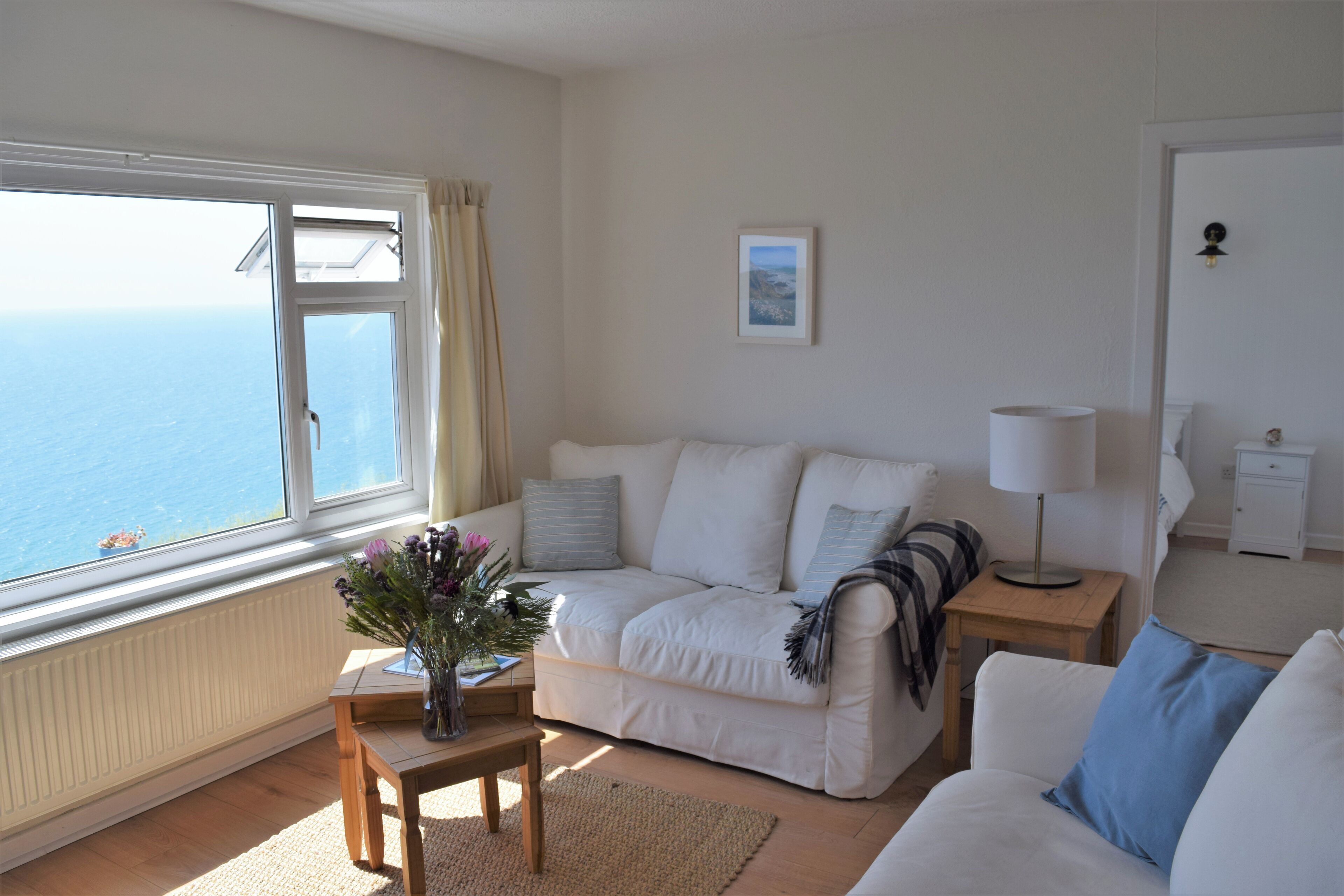 Un salon chaleureux avec un canapé, une petite table sur laquelle est posé un vase, et une vue sur la mer par une grande fenêtre.