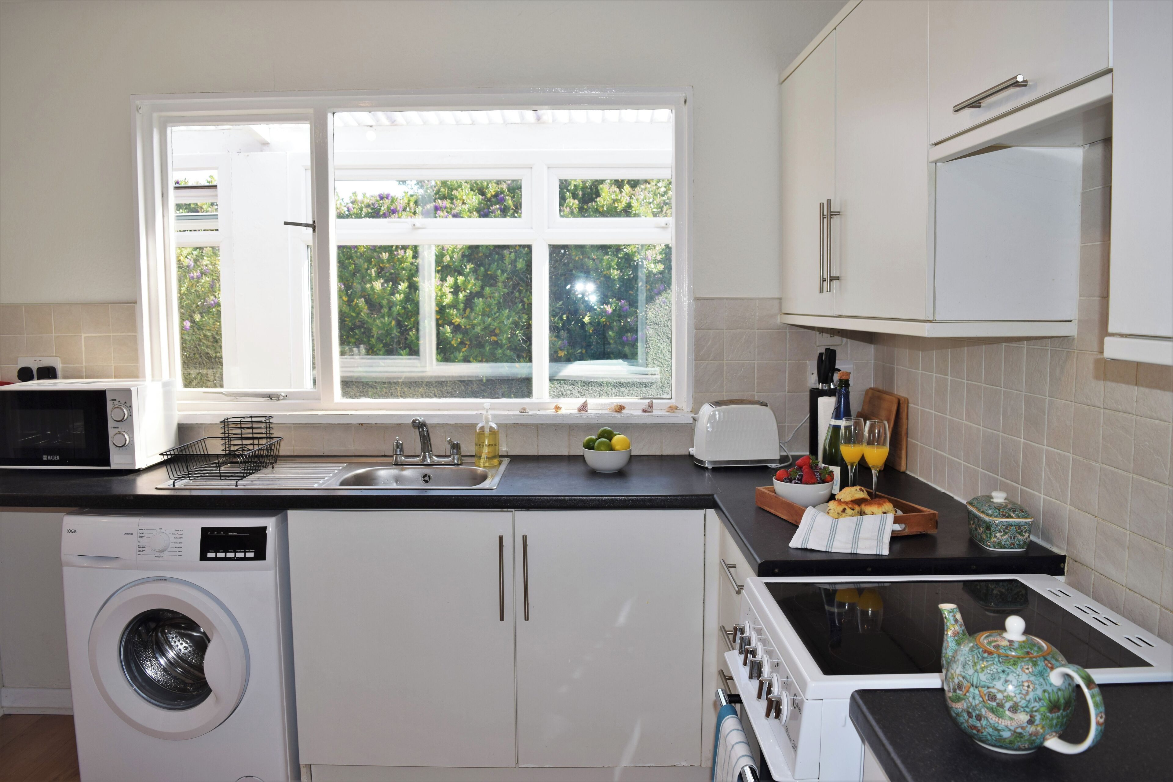 A modern kitchen with a washing machine, a sink, a window with a view of greenery, and a countertop with a teapot and other items.