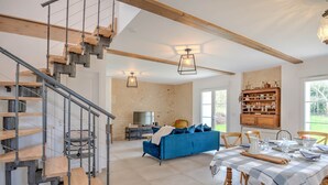 A modern living room with a blue sofa, wooden dining table, and a staircase with exposed beams.