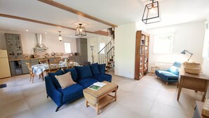 A spacious living room with a blue sofa, a wooden coffee table, and a dining area with a table and chairs. A staircase leads to an upper floor.