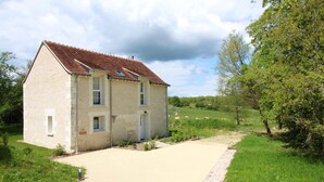 A two-story stone house with a tiled roof, a paved driveway, and a grassy field with trees and grazing animals in the background.