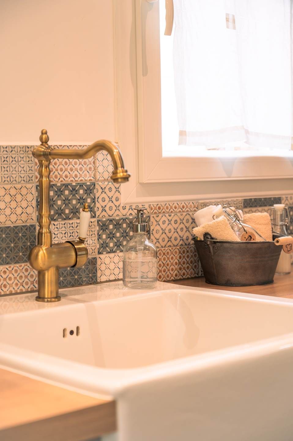 Un baño con un lavamanos blanco, un grifo de latón, azulejos estampados, una ventana con cortina y un cubo con toallas.