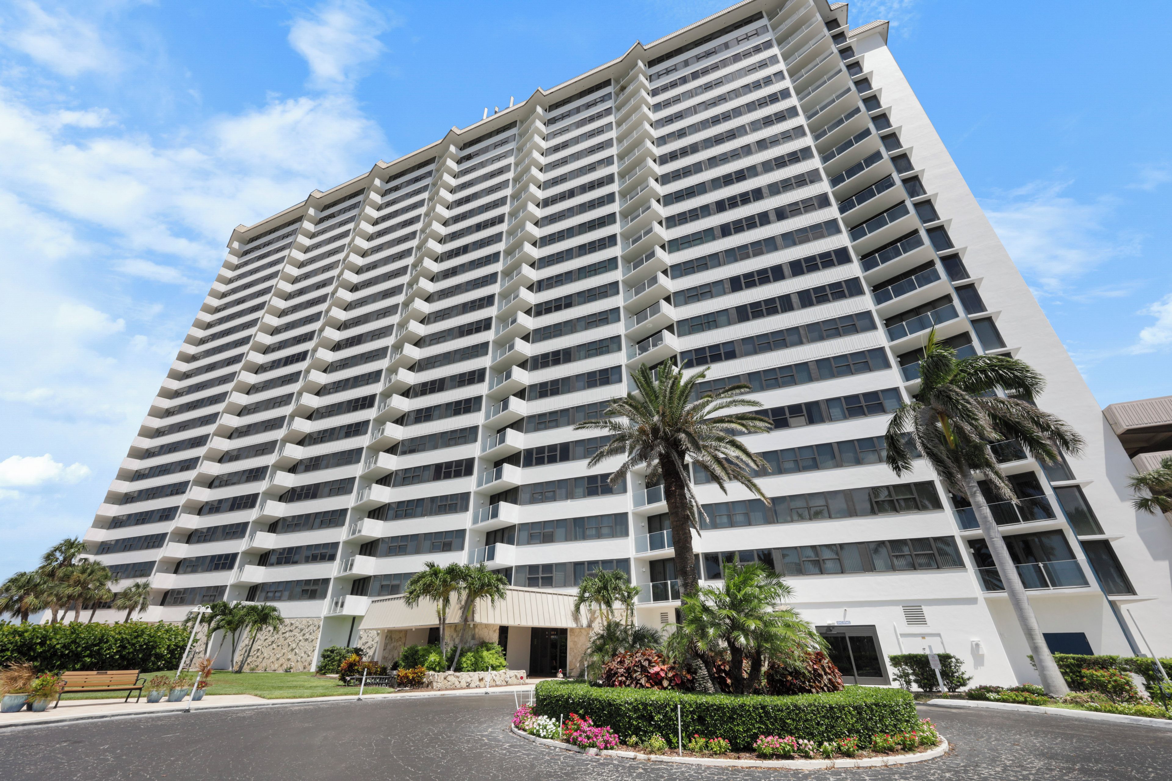 A high-rise hotel with multiple balconies, a landscaped courtyard with palm trees, and a clear blue sky.